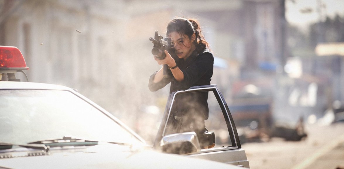 Sung-ae aims her weapon from the patrol car as destruction engulfs the village in Hope (2026).