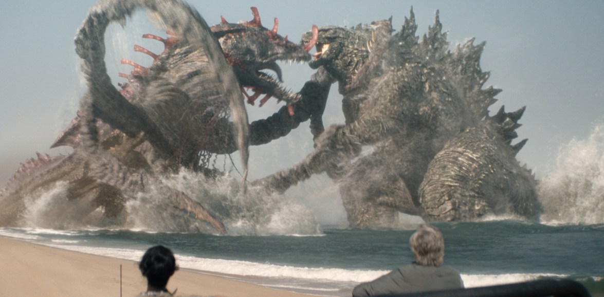 Godzilla and Titan X fight in the sea as two people observe from the beach, showing the scale of the monsters against the human story.